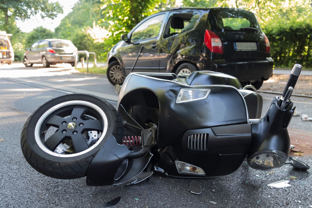 RoadAccidentWithMotorScooter | Detroit Personal Injury Attorneys Scooter accident scene with a damaged black scooter on its side, debris on the road, and a parked car in the background, illustrating the aftermath of a vehicle collision relevant to personal injury claims.