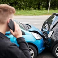 Person on phone assessing damage after car crash involving a blue and black vehicle, relevant to steps to take after an accident in Detroit.