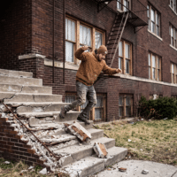 Man in a brown hoodie and beanie jumps off cracked concrete steps outside a brick apartment building, debris scattered around the stairs quickly.